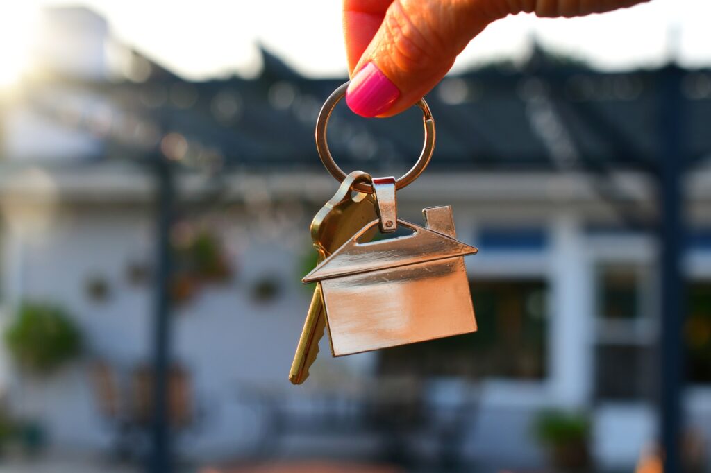 Female hand of realtor holding a set of keys outside home with pergola & flowers during golden hour.